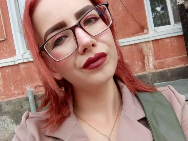 ValStephie has red hair, glasses, and red lipstick, with green eyes and a slight smile. She's wearing a pink jacket with a green strap. Her nose is pierced.