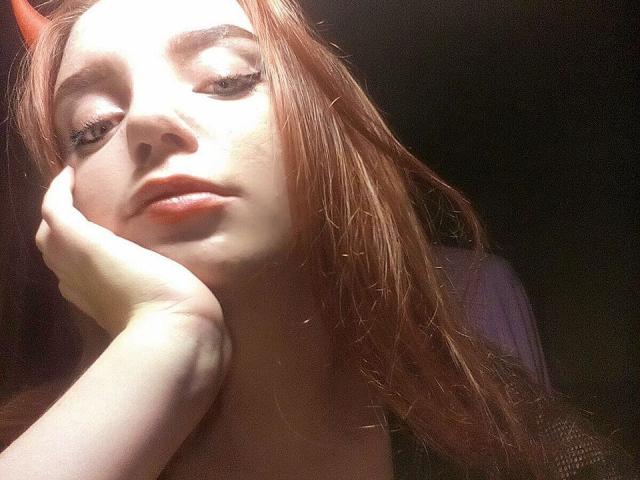 RedVelvett with pale skin, red horns, and long brown hair, sunlight highlighting her face, hand resting on cheek, slightly parted orange lips, looking bored, dark background.
