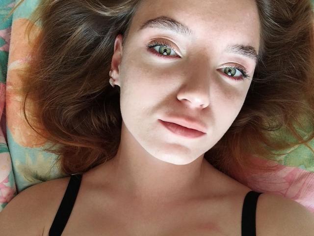RedVelvett has green eyes, light skin, and wavy brown hair. She's wearing black straps, with pink lips and small earrings. Her face is centered, looking at the camera.