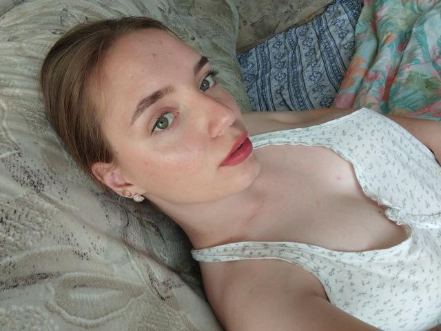 RedVelvett with light green eyes, red lips, and small tits in a white, floral-patterned tank top, lying on a bed with a patterned pillow. Blonde, fair-skinned.