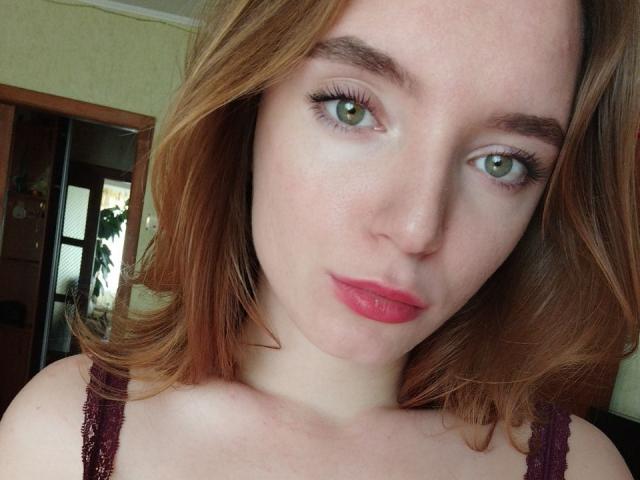 RedVelvett has green eyes, red lipstick, and light freckles, with wavy red hair, wearing a dark purple lace top, showing a cute and sexy face.