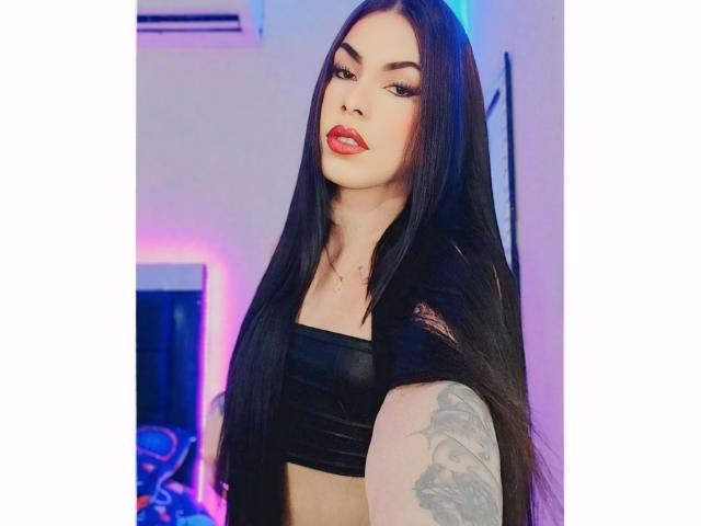 BriHotyX with long black hair, red lipstick, black strapless top, tattooed arm, looking at camera, medium tits, light skin, slightly parted red lips, sexy, confident, indoor, blue lighting.