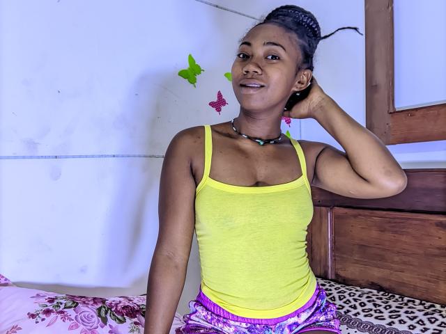 Elyanne with dark skin, small tits in a yellow tank top, purple and pink pants, braided hair, hand in hair, smiling, green and pink butterfly stickers on white wall.