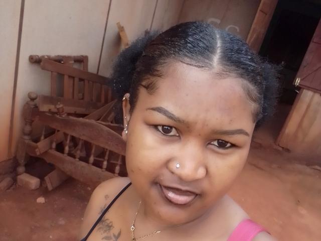 Marinahx has dark brown skin, black braided hair, a nose piercing, and a tattoo on her left shoulder. She's smiling with her tongue out, wearing a pink top.