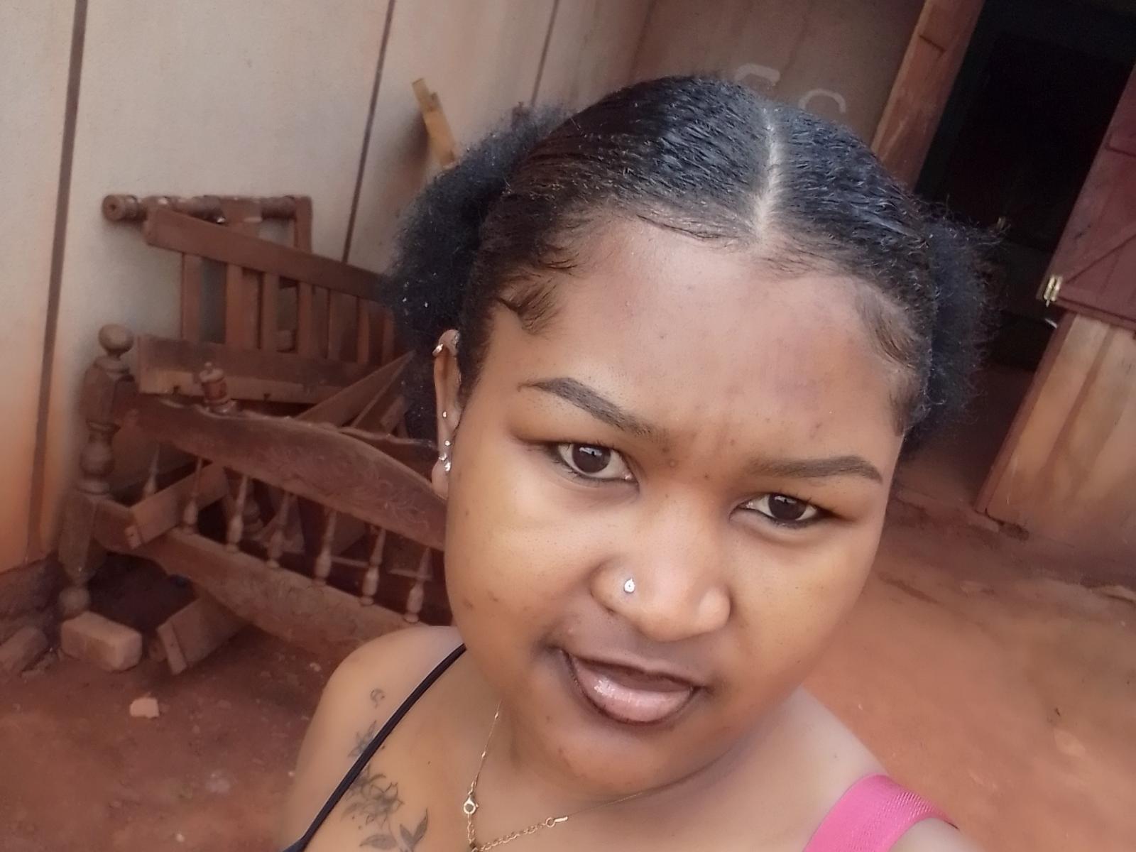 Marinahx has dark brown skin, black braided hair, a nose piercing, and a tattoo on her left shoulder. She's smiling with her tongue out, wearing a pink top.