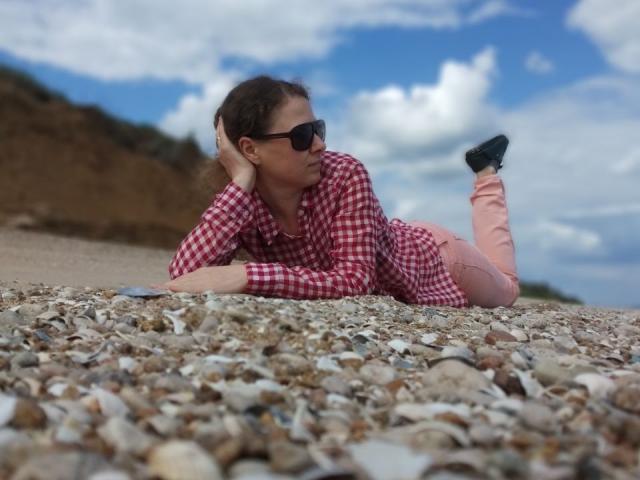 CurlyKsu, laying on pebble beach, wearing red check shirt, pink pants, black sunglasses, and black shoes, with head resting on hand, looking to the side, legs bent.