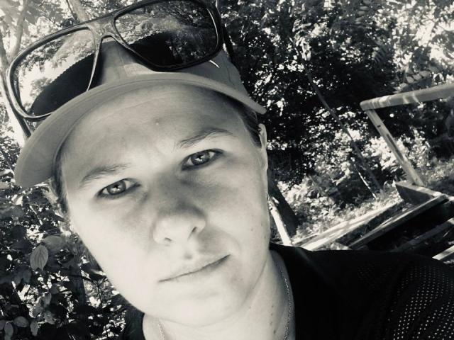 Black-and-white selfie of CurlyKsu with a serious expression, wearing a cap and sunglasses on their head, showing a slightly chubby face with visible pores and a small nose.