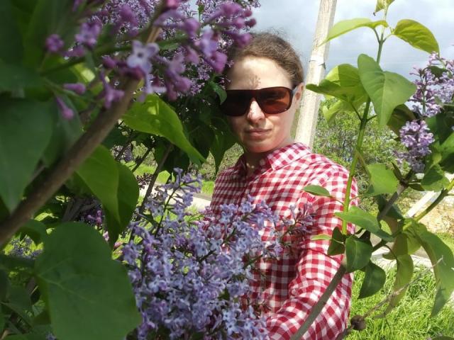 CurlyKsu with dark sunglasses, wearing a red checkered shirt, holding a cluster of purple lilacs, standing in a lush green garden, looking slightly to the side, smiling.