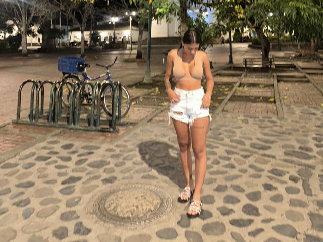 ThaliaAres with small boobs, tan skin, brown hair in a bun, wearing a beige bra and white shorts, standing on a stone-paved ground, looking down, in sandals.