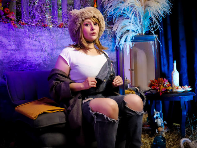DandelionQueen with brown hair, wearing a furry hat, white top, and ripped jeans, sits on a couch, removing a brown jacket, showing her knees and a cute face.