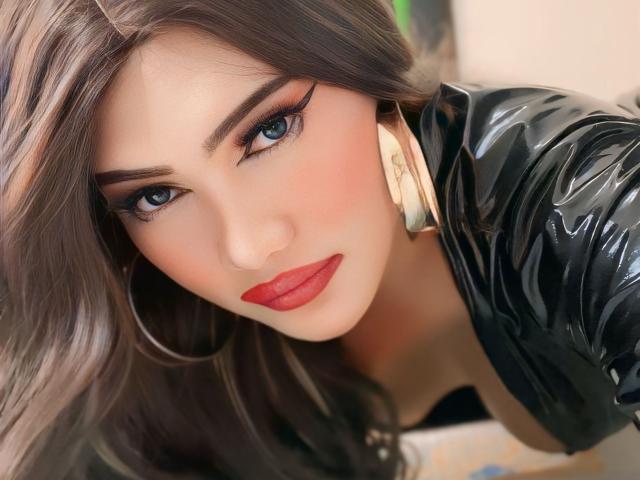 alwinamistress with red lips, blue eyes, and dark brown hair, wearing a black shiny top and gold earrings, looking seductively at the camera.