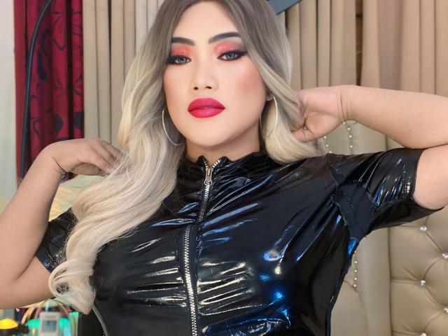 alwinamistress with long blonde hair, red lipstick, and black shiny latex top, hands in hair, wearing hoop earrings, looking at camera, confident, sexy, medium tits, buttoned top.