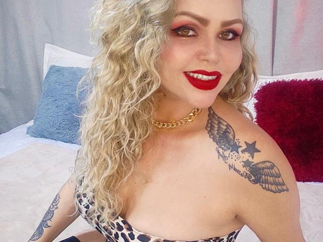 AnnaRichie with curly blonde hair, red lipstick, gold chain, leopard print top, tattoos on shoulder, smiling, showing tits, bed with blue and red pillows in background.