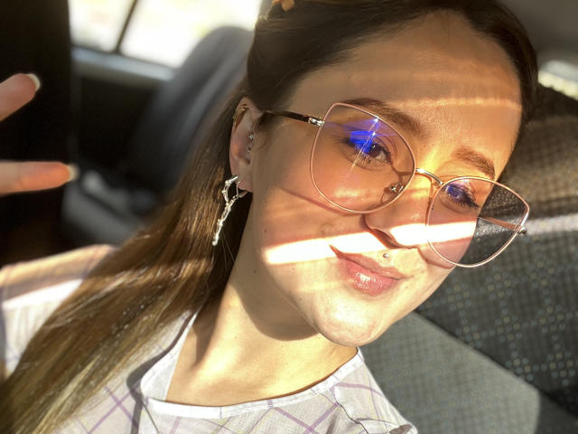 NinaBryons has light skin, brown hair, and wears big round glasses with blue reflection. She has a nose ring, lip ring, and cross earrings. Sunlight casts striped shadows.