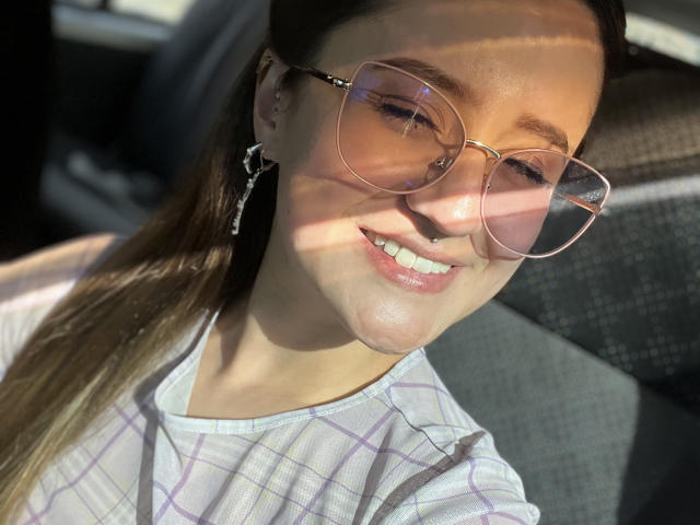 Nina Bryons, con cabello castaño claro, usando gafas de color rosa, sonriendo y mostrando dientes blancos, vestida con una camisa blanca a cuadros, con la luz del sol proyectando sombras en su rostro. Sus senos son ligeramente visibles..