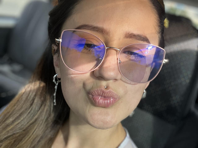 NinaBryons has puffy pink lips, a nose piercing, and wears pink aviator sunglasses with blue reflections. She's giving a pouty face, with brown hair in a ponytail.