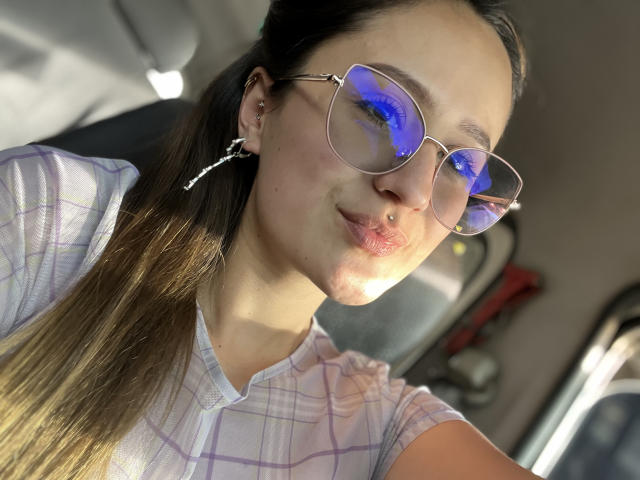 NinaBryons with puffy lips, wearing blue reflective glasses, and a pink checkered shirt, has brown hair in a ponytail, and small hoop earrings, sitting in a car.