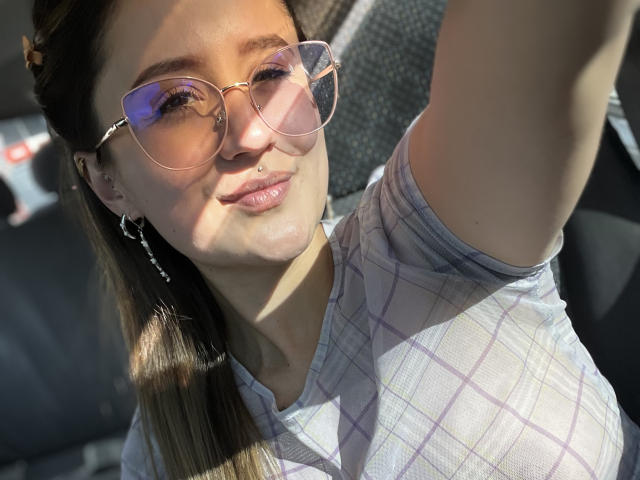 NinaBryons with pink-tinted glasses, plaid shirt, and nose ring, smiling in sunlight, showing her tits and a small silver earring, raising her arm.