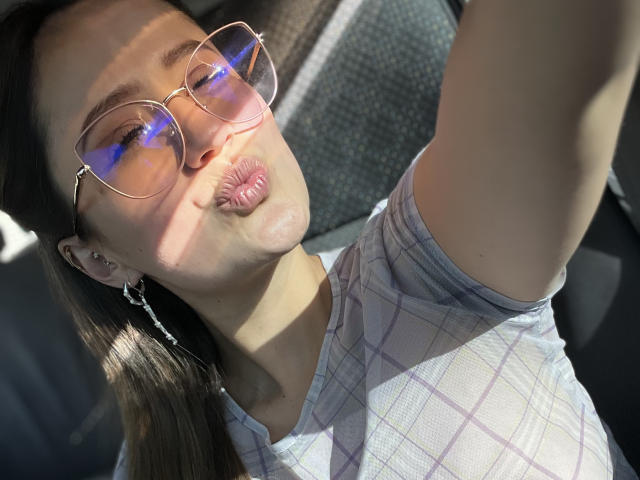NinaBryons with puffy lips, wearing glasses, plaid shirt, and earrings, taking a selfie, sunlight on her face, raising her right arm. Her brown hair is tied back.