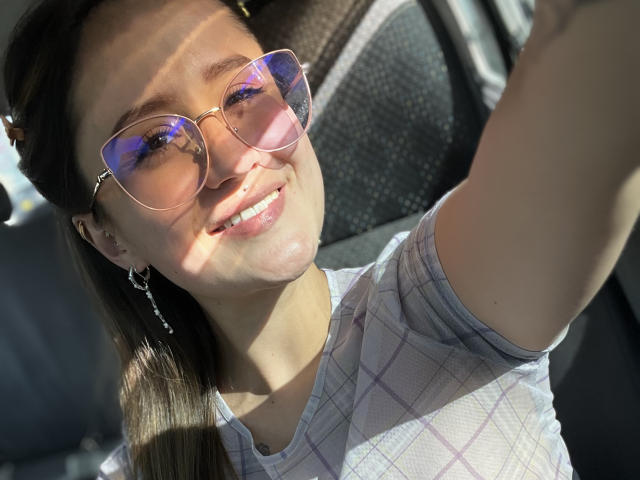 NinaBryons with pink reflective sunglasses, smiling, wearing a white plaid shirt, sunlight in her brown hair, small earrings, taking a selfie in a car.