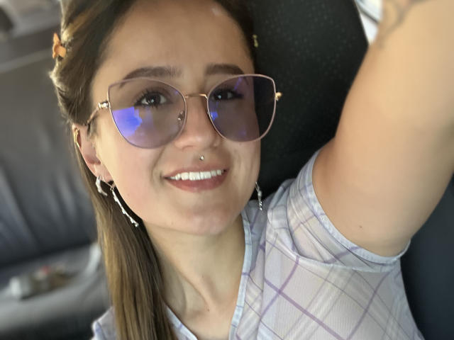 NinaBryons with brown hair in a ponytail, wearing pink-tinted aviator sunglasses, a plaid shirt, and a nose ring, smiling at the camera in a car.