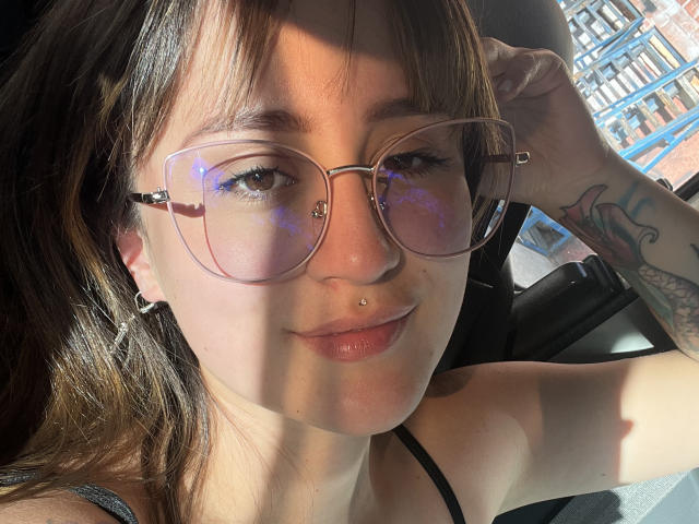 NinaBryons with glasses, nose ring, and lip piercing, sunlight on her face, arm tattoo, brown hair, slight smile, car seat, reflective blue light on glasses.