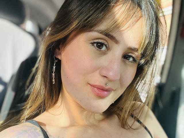 NinaBryons has brown eyes, a nose piercing, a lip piercing, and long brown hair with bangs, wearing a black top, with earrings, sitting in a car.