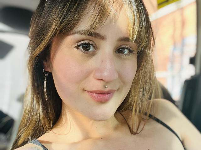 NinaBryons has light brown hair, a nose piercing, and a slight smile. She's wearing a cross earring and a black top, with her brown eyes looking forward.