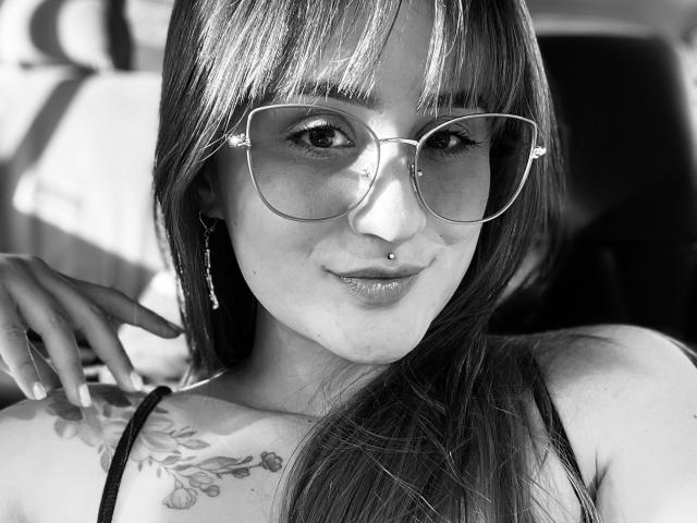 NinaBryons with long hair, wearing large glasses, nose piercing, and a tank top, showing a tattoo on her right shoulder, looking at the camera with a slight smile.