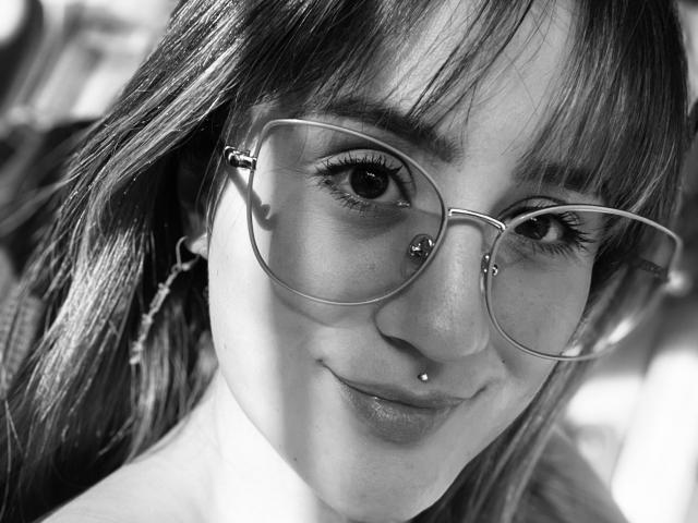 NinaBryons with glasses, nose piercing, and lip piercing, smiling slightly, showing off her cute face and freckles, in a black-and-white photo. Her ass is not visible.