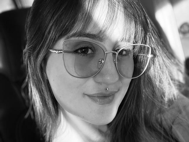 black-and-white photo of NinaBryons with glasses, septum piercing, and a slight smile, showing her ass-chin and subtle makeup, sitting in a car, looking forward.
