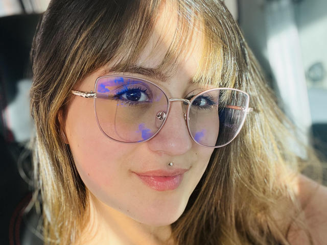 NinaBryons has light brown hair, blue eyes, and wears clear glasses with purple smudges. She has a nose ring and a lip piercing, smiling softly.