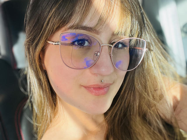 NinaBryons has blue eyeshadow, pink-framed glasses, a nose piercing, and light brown hair with bangs. Her pink lips are slightly parted. She's wearing a black shirt.
