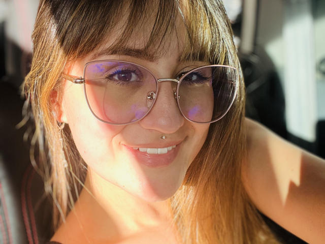 NinaBryons with light brown hair, smiling, wearing rose-tinted glasses, small nose ring, and pink lipstick, sunlight on her face, hand in car, wearing no top.