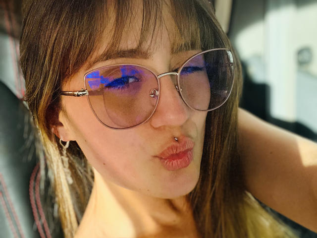 NinaBryons with pouty lips, nose piercing, and brown aviator sunglasses reflecting blue and purple, has a small hoop nose ring and wears a thin hoop earring.