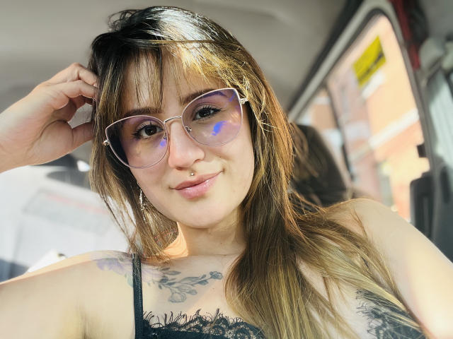 NinaBryons with glasses, smiling, has brown hair, tattoos on chest, black lace top, nose ring, sun reflection in glasses, hand in hair, car interior.