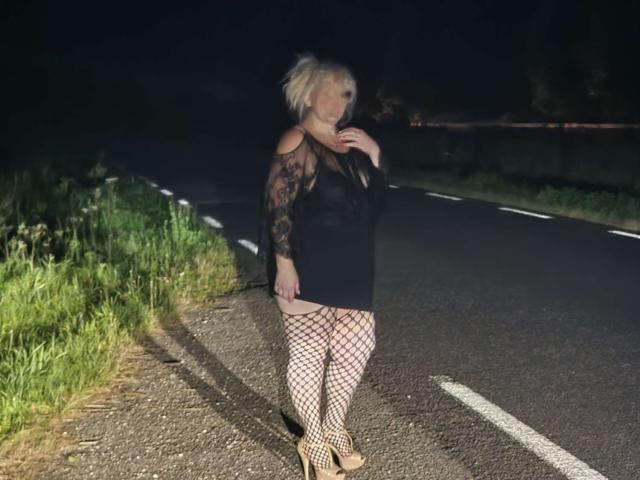 Enchaleur69X with blonde hair, black lace dress, fishnet stockings, and high heels, standing on a dark road at night, her hand near her chin, showing her big ass.