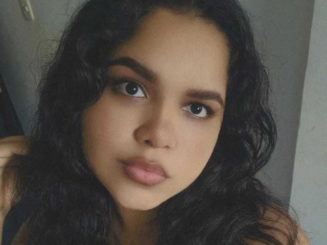 MillySofia25 has dark, wavy hair, brown eyes, and pinkish lips, with a neutral expression, and a light brown skin tone, looking straight into the camera.