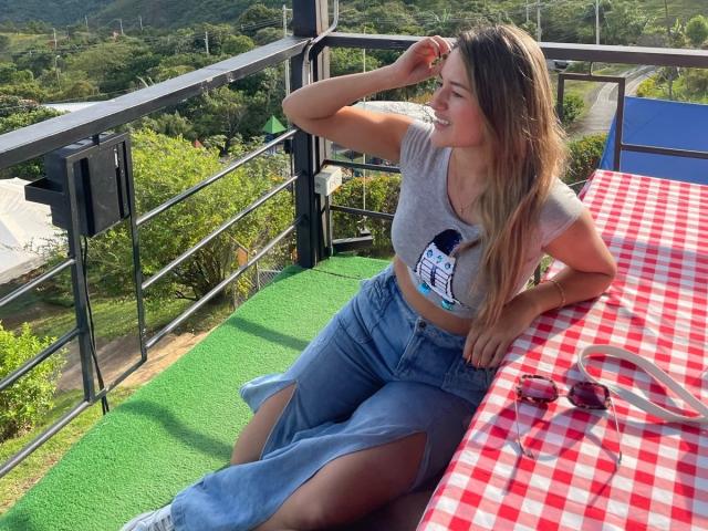 MariaVani with long blonde hair, gray crop top, and blue jeans, sitting on green turf, leaning on red checkered table, smiling, sunglasses on table, mountains in background.