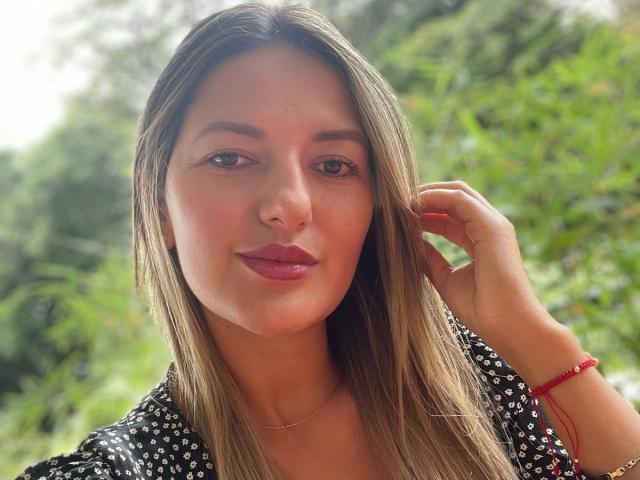 MariaVani has long brown hair, fair skin, and pink lipstick, wearing a black floral dress, with a red bracelet on her right wrist, in a green, blurry background.