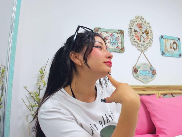 Genesiss with black hair in a ponytail, wearing a white shirt, has her finger on her chin, red lipstick, and black sunglasses on her head, looking to the side.