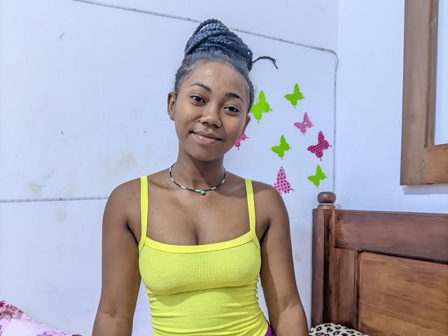 Elyanne has dark skin, small breasts, and wears a yellow tank top with a beaded necklace. She has gray braided hair and a slight smile, standing by a wooden bed.