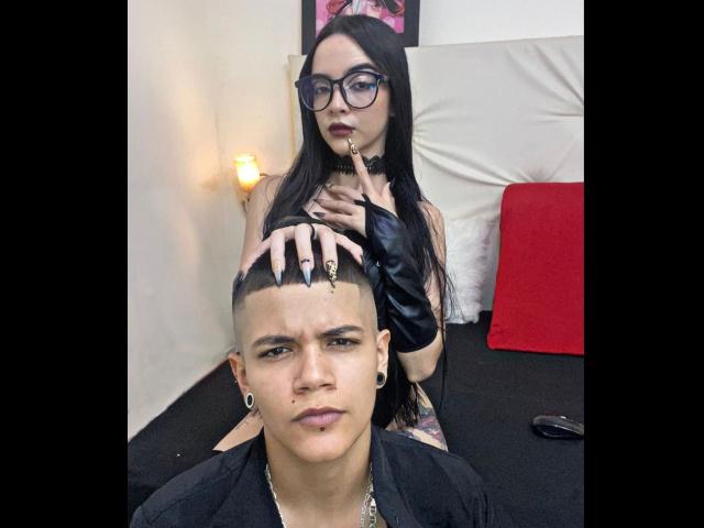 EmilyAndSam with black hair, glasses, and goth makeup, finger in mouth, standing behind a bald, tatted, pierced, and dressed-in-black guy with a serious face.