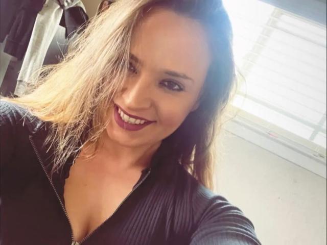 AshsLeezy69 has long blonde hair, smiling with dark purple lipstick, wearing a black zip-up top, showing cleavage, and has light skin, looking cute and sexy.