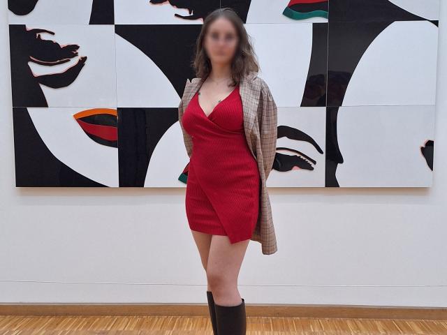 CoralieLaCoquine with blurred face, wearing a red dress with plunging neckline, plaid jacket, and black boots, standing in front of black-and-white pop art wall. Boobs visible.