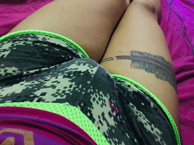 ArianaUnicorn's ass in pixel-patterned gym shorts with neon green trim, showing a tattoo on the right thigh, lying on a pink bed.