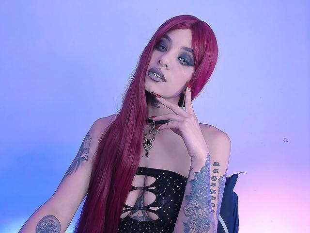 DarkAngeliq con cabello rojo largo, sombra de ojos azul y tatuajes, vestida con un vestido negro sin tirantes con cierre frontal de cordones, tocándose la barbilla, mirando a la cámara, piel pálida..