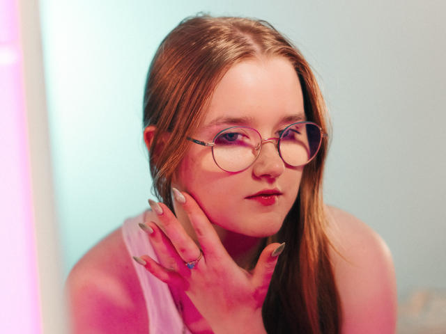 BonnieBell with pink glasses, long brown hair, pink lipstick, and a white sleeveless top, touching her face with manicured nails, looking slightly to the side.