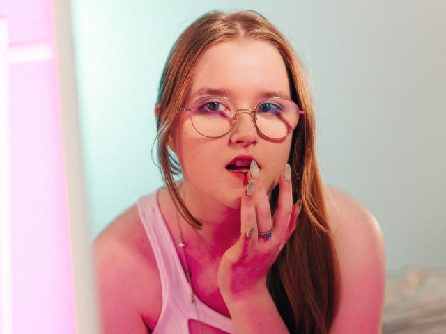 BonnieBell with blonde hair, blue eyes, and glasses, touching her slightly open pink-liped mouth with her finger, wearing a pink tank top, pink neon light on left.