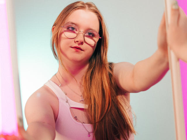BonnieBell with long red hair, glasses, and pink tank top, showing off her tits with a confident smile, holding a mirror. Her hand is on the right.